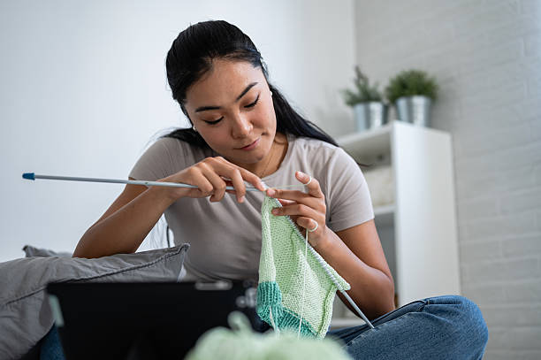 A young woman sits comfortably at home, focused and content while knitting a green and white scarf, engaging in a calming self-care activity.