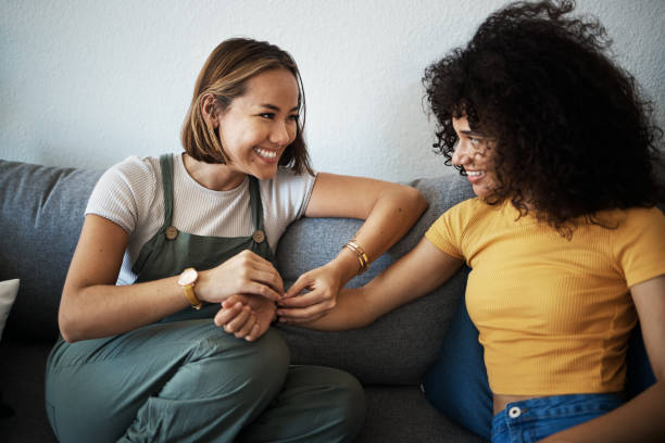 Two women sitting on a couch, smiling and engaging in a warm, supportive conversation. One woman is gently holding the other’s hand, conveying emotional connection and open communication in a relaxed setting.