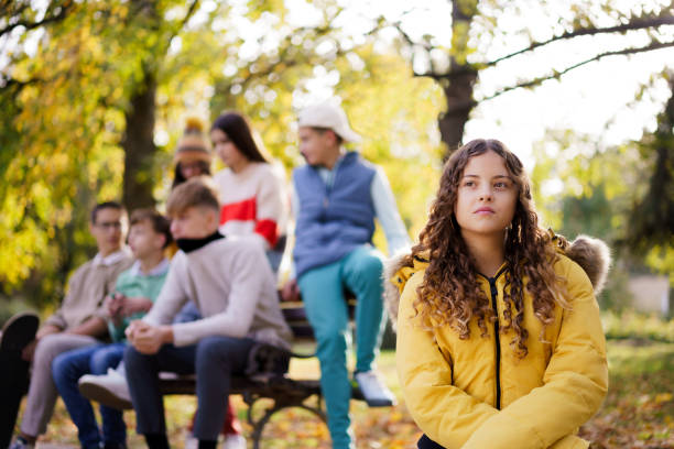 A teenage girl in a yellow jacket sitting alone on a park bench, looking sad and isolated while a group of teens socializes in the background. The setting is outdoors in autumn.