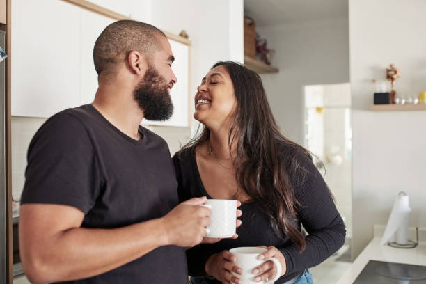 Happy couple sharing a light moment in the kitchen while holding coffee mugs, representing intimacy, joy, and emotional connection before marriage.