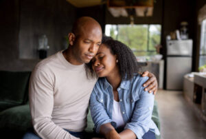 A Black couple sits closely on a couch in a cozy living room, with the woman leaning her head on the man’s shoulder and his arm wrapped around her in a comforting embrace. They appear to be sharing an emotional moment.