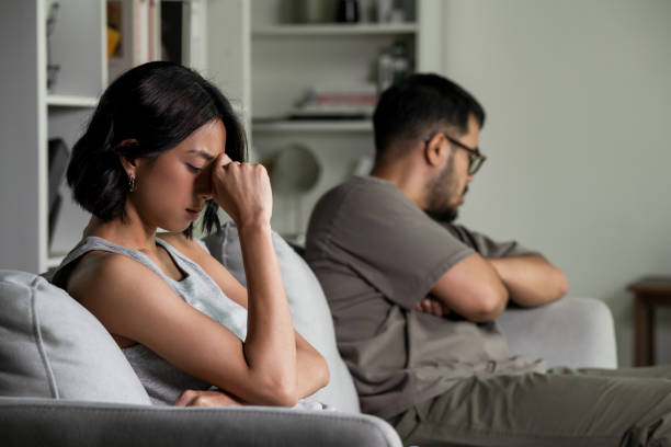 An Asian couple sits apart on a couch, both looking upset and distant. The woman holds her head in her hand in frustration while the man looks away with his arms crossed. Tension and emotional disconnection are visible.