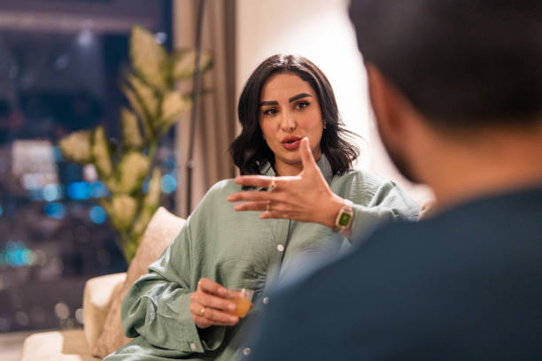 A Middle Eastern woman gestures expressively while speaking during a serious conversation with a man whose back is to the camera. The setting appears to be an evening living room discussion, with city lights in the background.