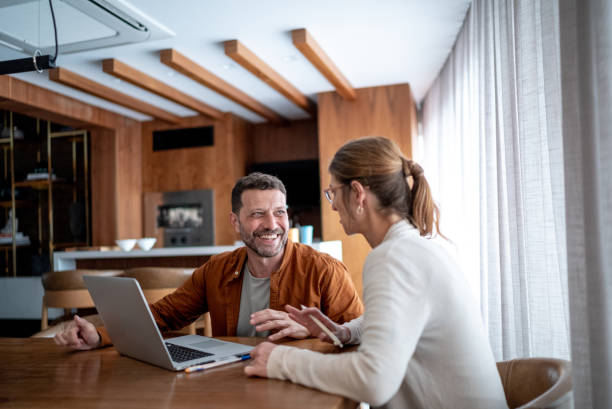A smiling man and woman sitting at a table with a laptop, engaged in a collaborative and positive conversation in a modern home office setting.