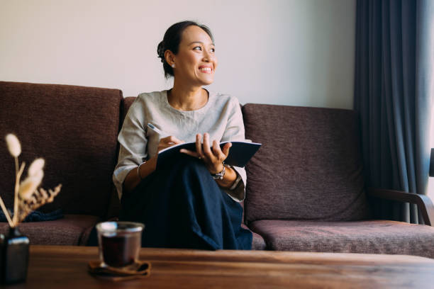 Young Asian woman sitting comfortably on the couch in her living room, writing in her notebook.
