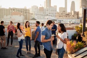 Young adults socializing at a rooftop party during sunset in the city, with string lights and drinks creating a lively, casual atmosphere.