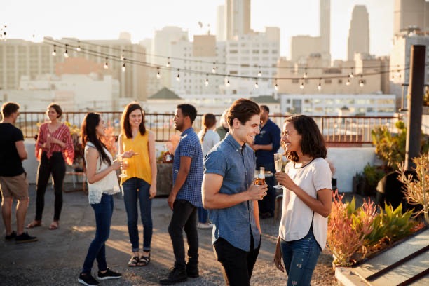 Young adults socializing at a rooftop party during sunset in the city, with string lights and drinks creating a lively, casual atmosphere.