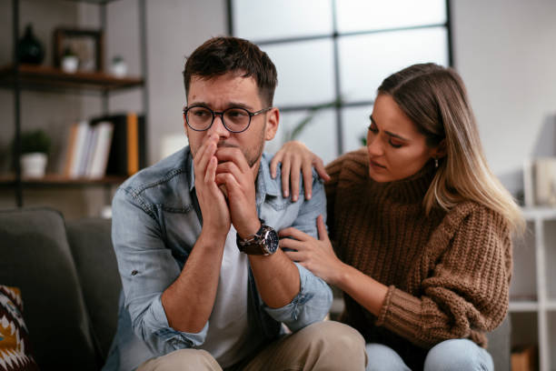 A woman gently places her hands on a distressed man's shoulder, offering comfort and support as he sits on a couch, deep in thought. The setting appears to be a home or therapy-like environment.