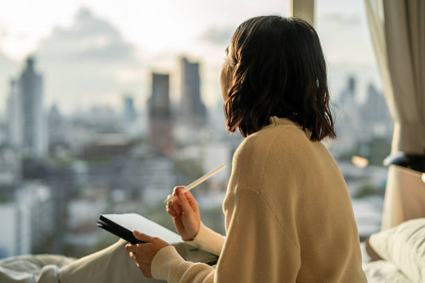 A woman sits on a bed holding a digital tablet and stylus, gazing out a window at a city skyline during sunrise or sunset, lost in thought.