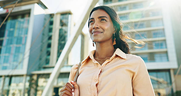 A confident young woman stands outside in front of modern buildings, smiling slightly as sunlight shines on her face. She appears empowered and thoughtful, dressed in a peach button-up shirt.