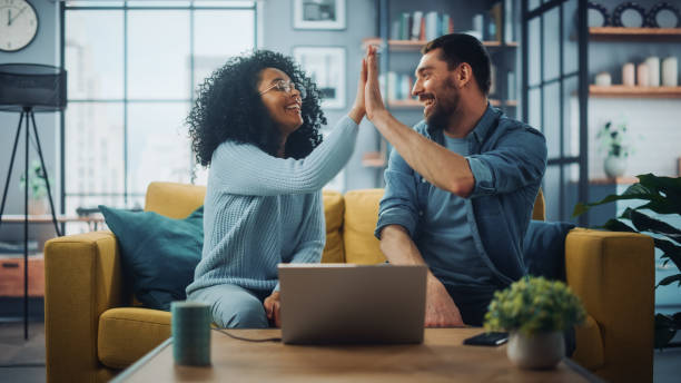 A cheerful couple sits on a sofa in a cozy living room, high-fiving each other with a laptop on the table in front of them.