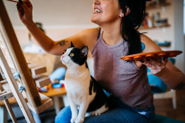 A woman laughing as she paints on a canvas, with a black-and-white cat sitting on her lap, both facing the artwork.