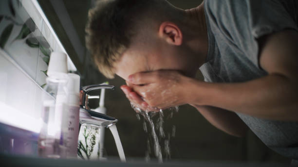 Caucasian boy comes to sink in bathroom, washes face with cold water and looks at his reflection in the mirror. Teenager stands in front of basin and makes morning bathroom routine. Lifestyle concept.