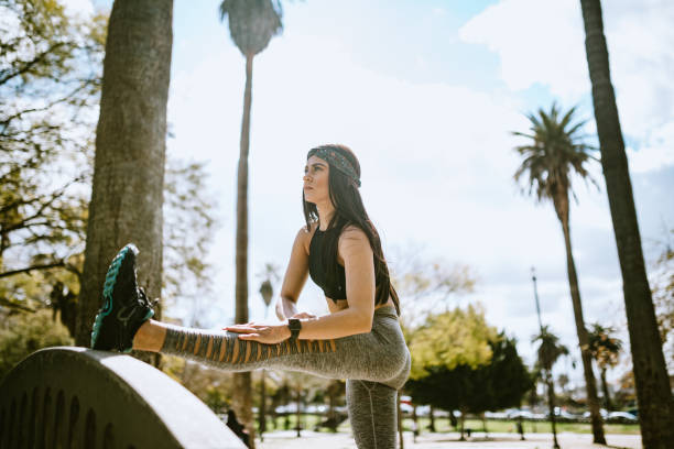 A fit young woman in athletic clothing stretches her leg on a park bridge railing, surrounded by palm trees and sunny skies, suggesting a healthy and mindful lifestyle.