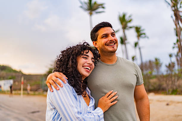 A happy couple stands arm-in-arm on a tropical beach, smiling and looking into the distance with palm trees in the background.