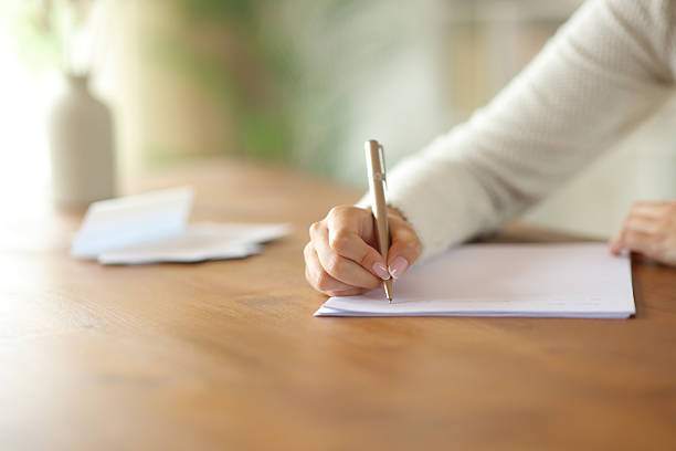 Close-up of a woman’s hand writing on a piece of paper at a wooden desk, with soft focus on the background.