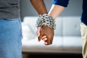 Two people holding hands tightly wrapped in metal chains, symbolizing emotional entrapment or codependency in a relationship. The background is blurred, drawing focus to the symbolic gesture.