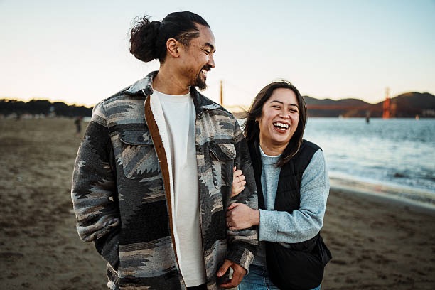 A smiling couple walks together on the beach during sunset, laughing and enjoying each other’s company, with the Golden Gate Bridge in the background.