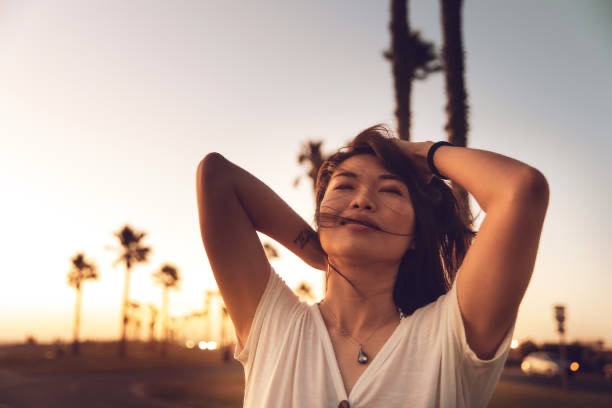 A young woman smiling with her eyes closed, standing outdoors at sunset with palm trees and the breeze blowing her hair.