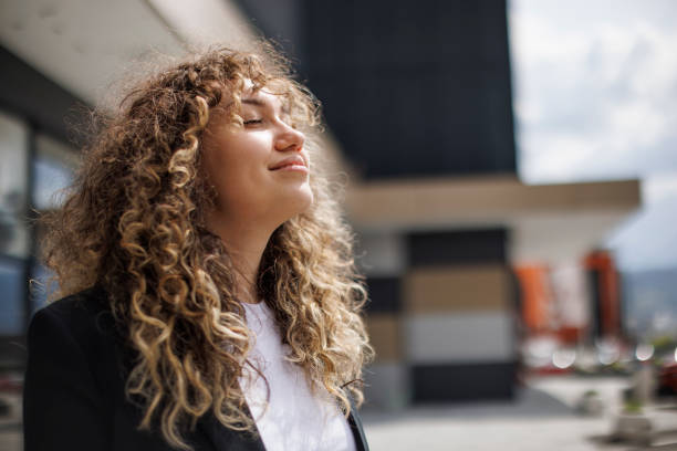 A curly-haired woman standing outside in the sun with her eyes closed and a peaceful smile, wearing a black jacket and white shirt.