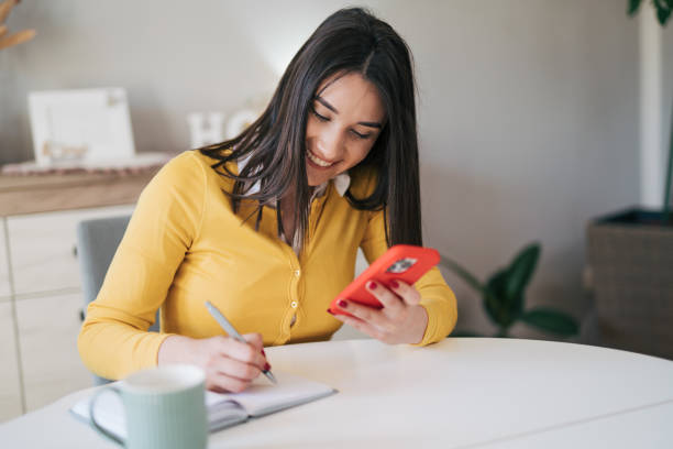 Young woman in a yellow sweater writing in a notebook while holding a red smartphone, focused and cheerful.