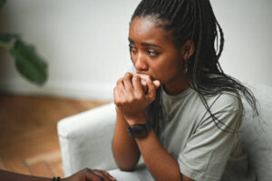Young Black woman sitting in a chair, looking anxious and contemplative, holding her hands near her mouth during a therapy session.