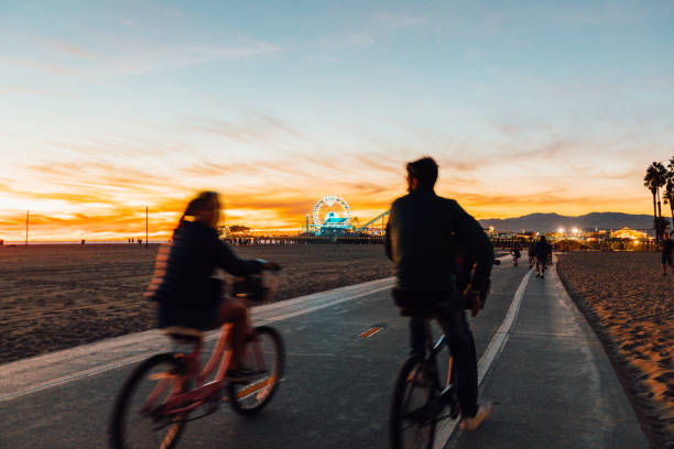 Two people ride bikes along a paved beach path at sunset, with the Santa Monica Pier and Ferris wheel glowing in the background.