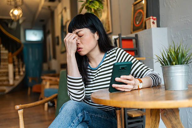 An Asian woman sitting in a café, holding her phone in one hand and rubbing her forehead with the other, appearing frustrated or stressed.