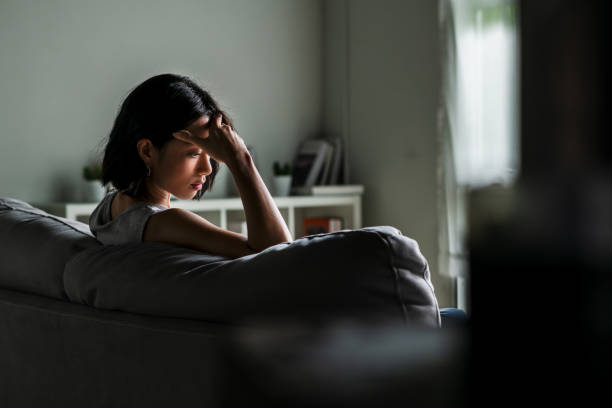 Woman sitting on a couch in a dimly lit room, resting her head in her hand, appearing stressed or deep in thought.