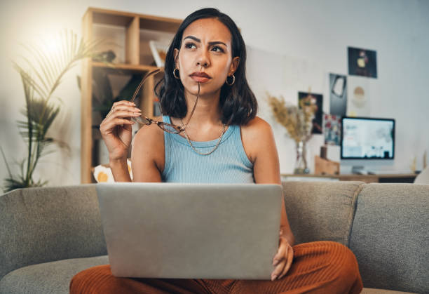 A thoughtful woman sitting on a couch with a laptop on her lap, holding her glasses and looking upward as if reflecting or brainstorming.