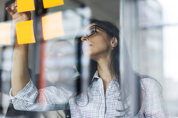Professional woman planning ideas with sticky notes on a glass wall, focused and thoughtful in a modern workspace.