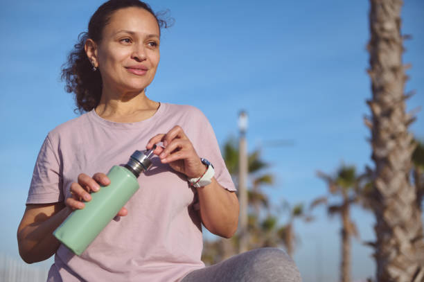 Smiling woman outdoors in athletic wear holding a green water bottle, with palm trees and blue sky in the background, suggesting a healthy lifestyle.