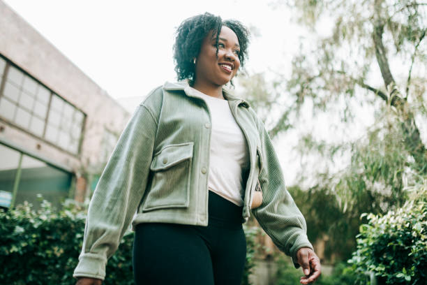 A smiling young Black woman walking outdoors, wearing a green corduroy jacket and black pants, surrounded by greenery and urban buildings.