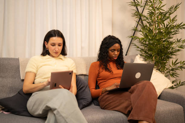 Two women sitting on a couch, working side-by-side with a tablet and a laptop, focused and collaborative.