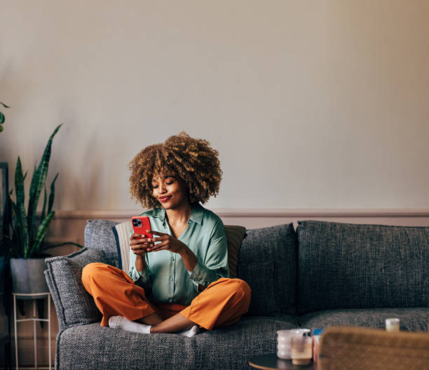 Young woman with curly hair sits cross-legged on a gray couch, smiling as she looks at her phone.