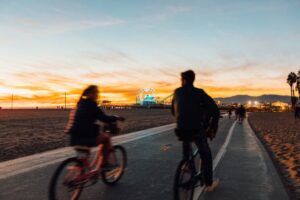 Two people ride bikes along a paved beach path at sunset, with the Santa Monica Pier and Ferris wheel glowing in the background.