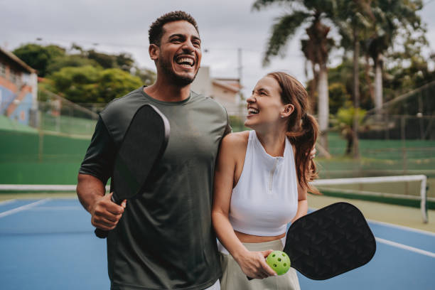 A smiling couple walks arm in arm on a pickleball court, holding paddles and a ball, enjoying a lighthearted moment outdoors.