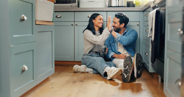 A happy couple sits on the kitchen floor laughing and giving each other a high five, celebrating a joyful moment together.
