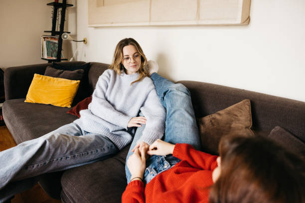 Two women sit comfortably on a couch, one listening attentively while the other lies down, suggesting a calm and supportive conversation.