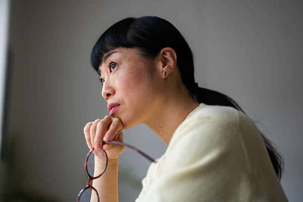 Woman holding her glasses and looking out a window, appearing contemplative and emotionally reflective.