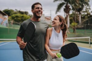 A smiling couple walks arm in arm on a pickleball court, holding paddles and a ball, enjoying a lighthearted moment outdoors.
