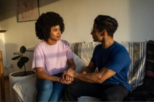 Couple sitting on a couch holding hands while having a calm and supportive conversation.
