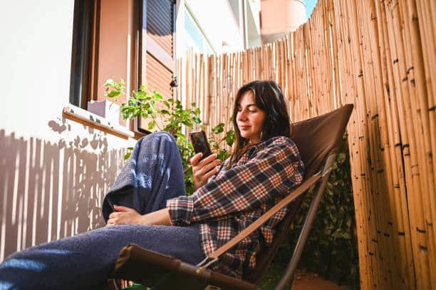 Woman sitting outside in a chair using her phone, appearing relaxed and taking a break from productivity and perfectionistic pressure.
