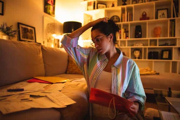 Woman sitting on a couch surrounded by papers, looking overwhelmed and stressed due to perfectionism and high expectations.
