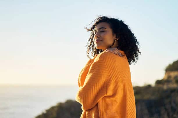 Woman standing outdoors near the ocean with a peaceful expression, representing self-acceptance and letting go of perfectionism.