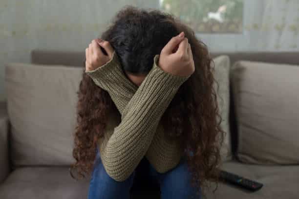Young girl sitting on a couch with her face covered, appearing distressed or overwhelmed by emotions.