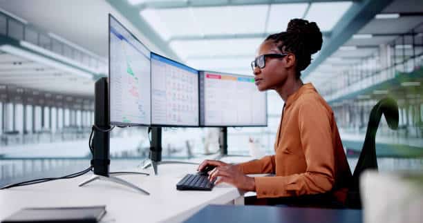 Professional woman focused on work at a desk with multiple computer monitors in a modern office environment.