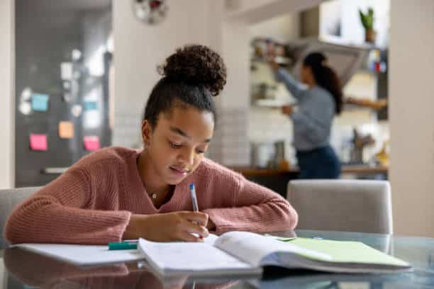 Young girl focused on completing schoolwork at home while a parent works in the background.