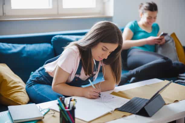 Teen girl studying at home, taking notes and completing homework while managing distractions in a shared living space.