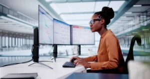 Professional woman focused on work at a desk with multiple computer monitors in a modern office environment.